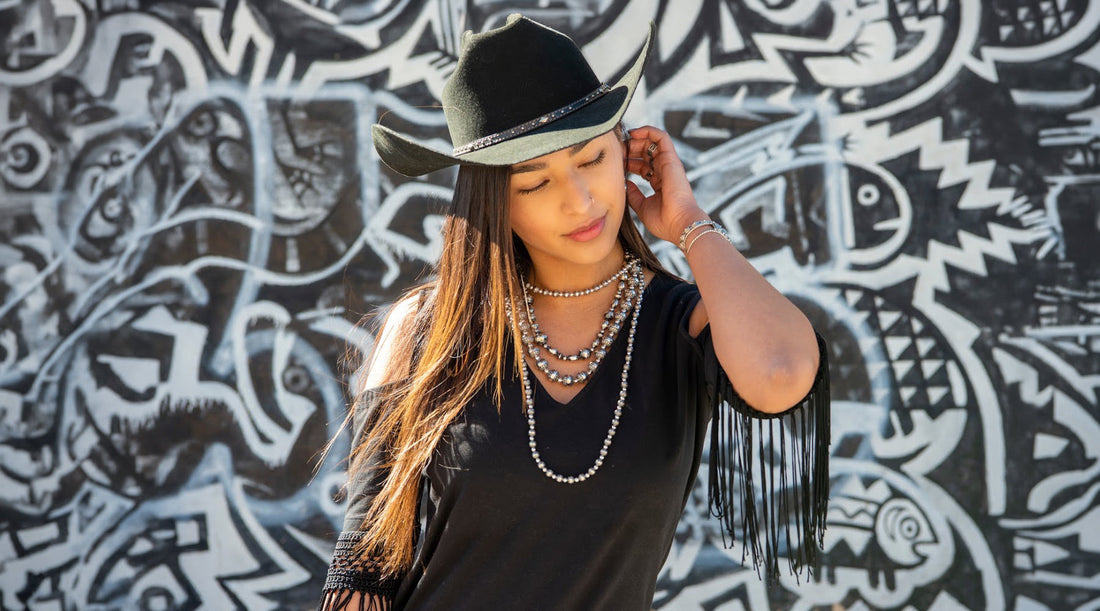Woman wearing American West Jewelry layered sterling silver Native pearl bead necklaces, ring, and bracelet styled in a black fringe top and cowboy hat against a mural, promoting Santa Fe Indian Market
